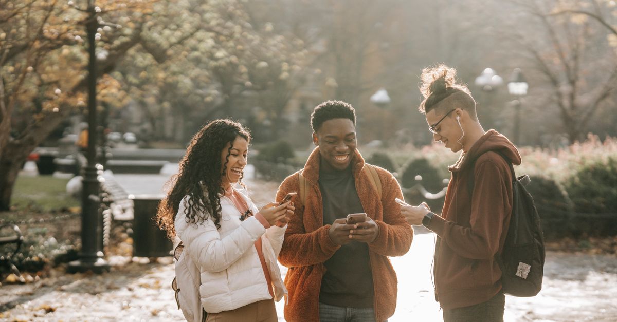Delighted young diverse group of male and female students with backpack standing in city park on sunny autumn day and surfing modern smartphones
